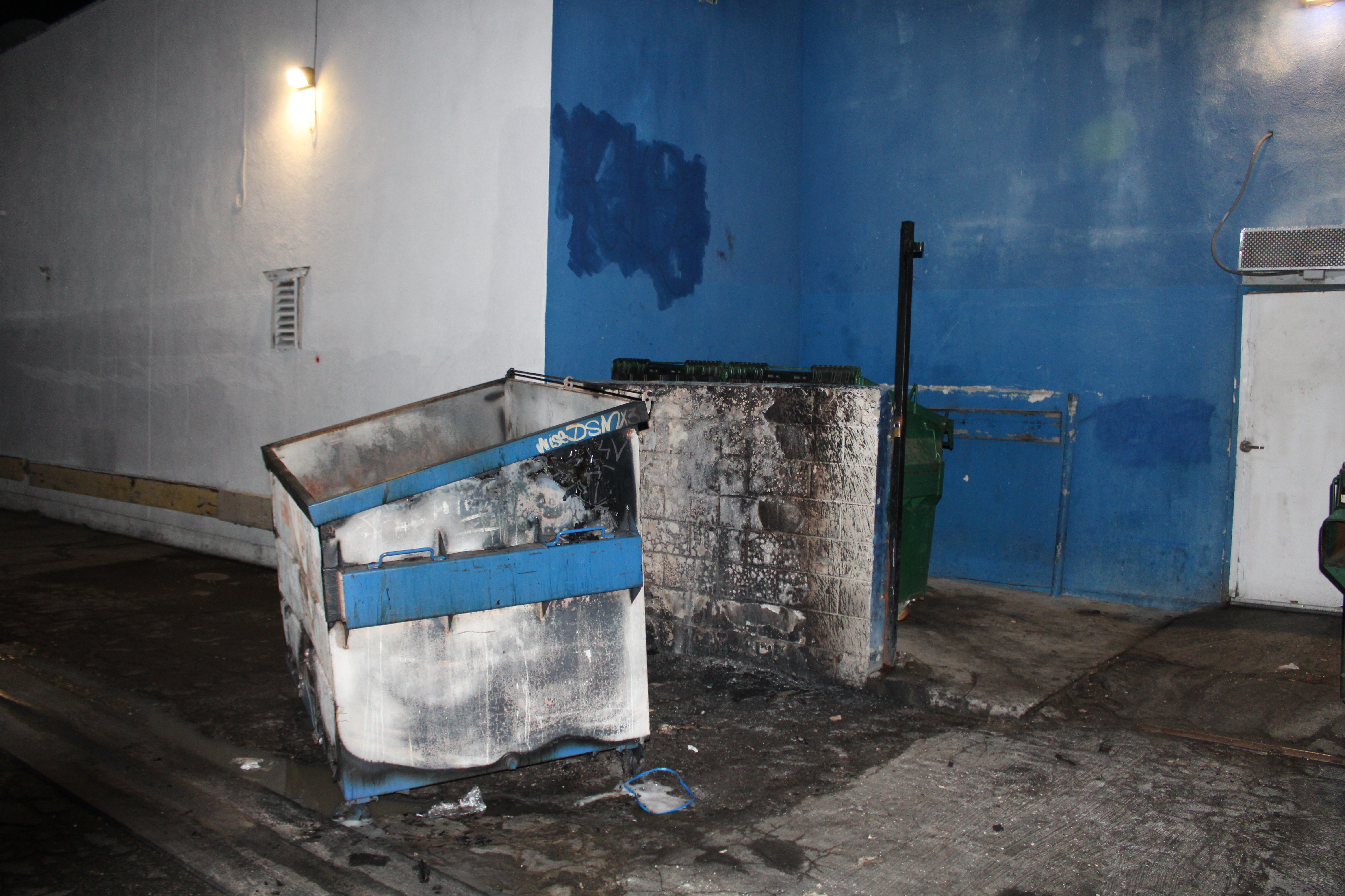 Dumpster behind a commercial building with black marks and melted paint on both, from a previous fire.