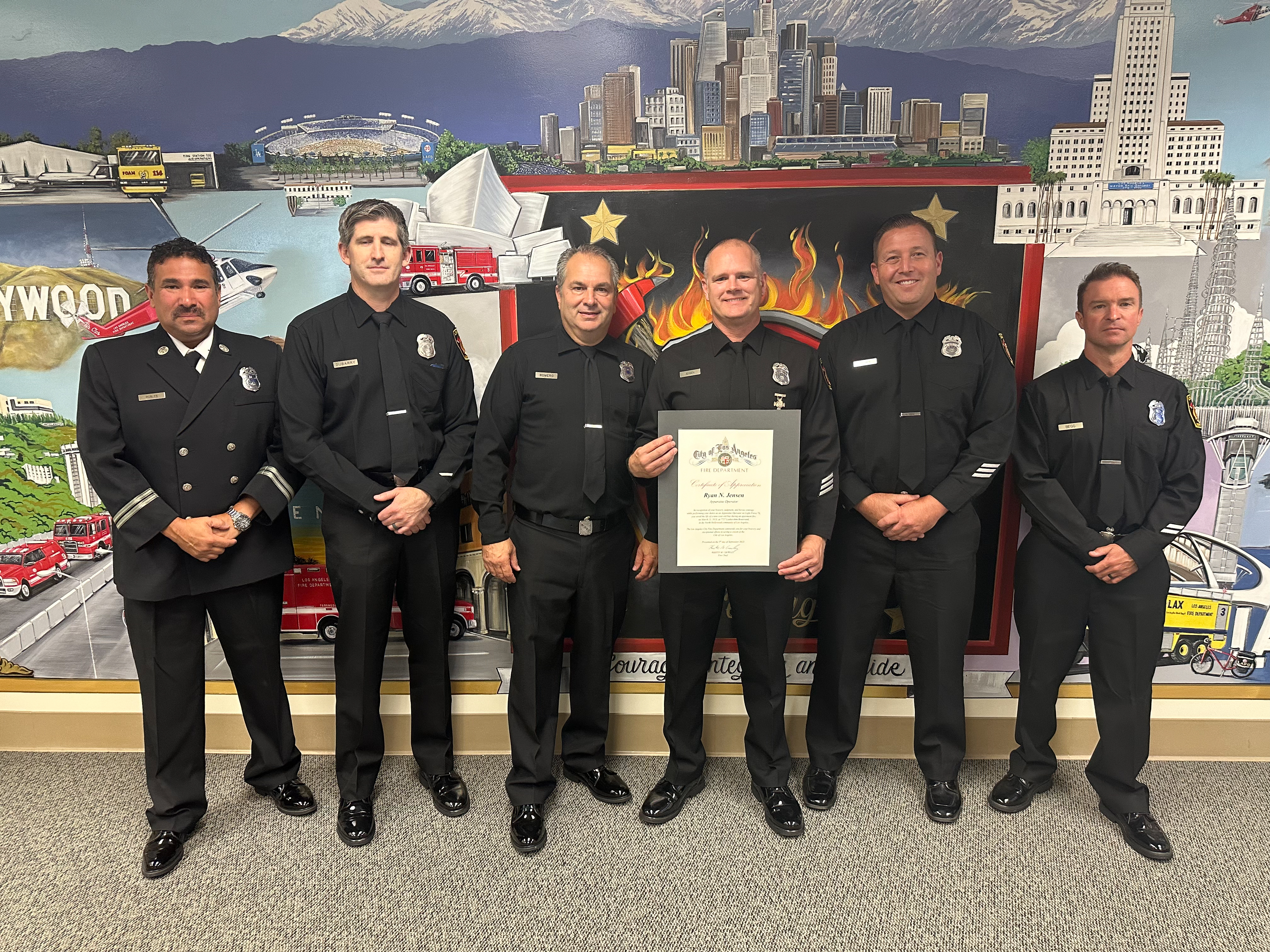 Six firefighters in dress uniforms standing together, the one in the center is holding a certificate.